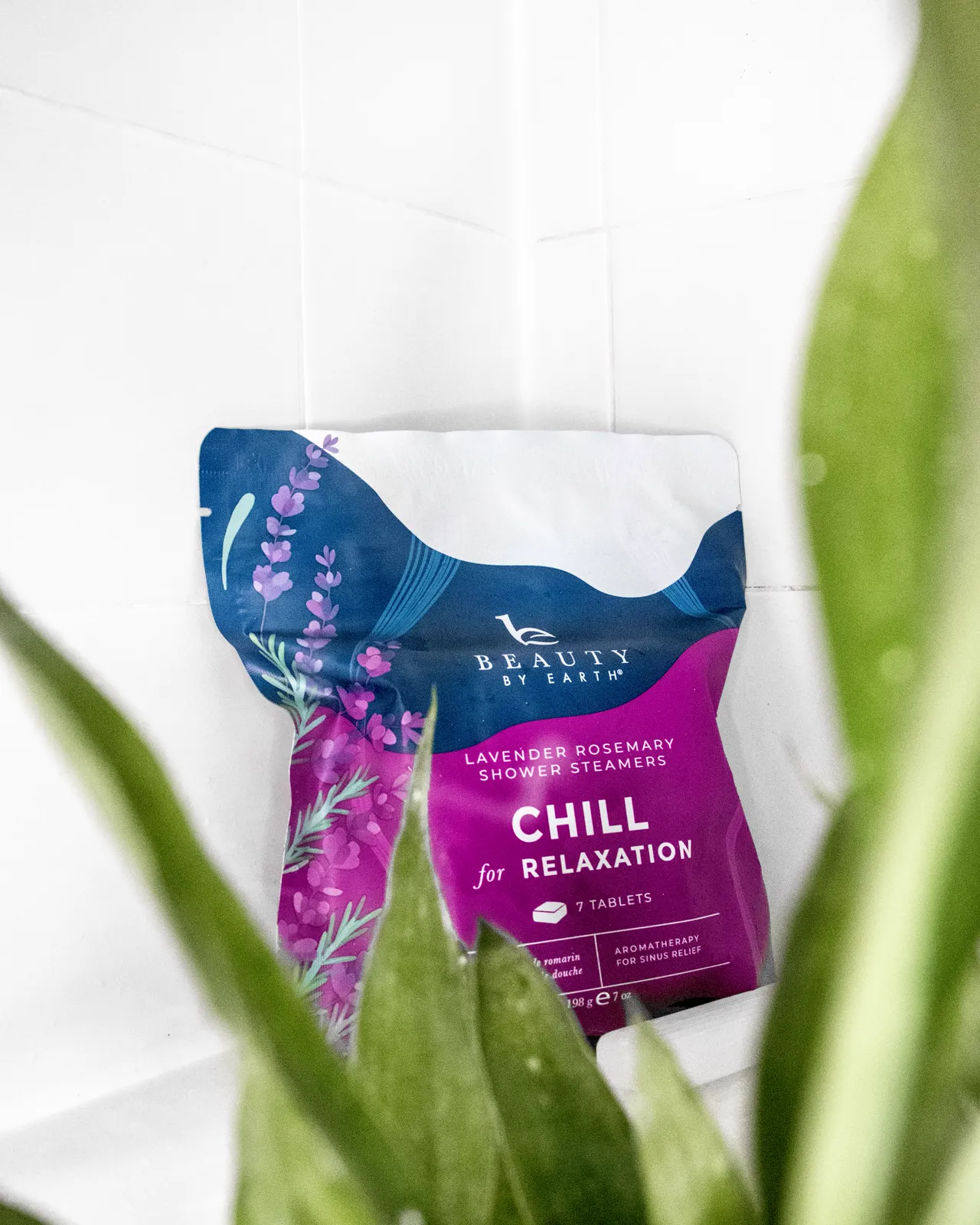 A Beauty by Earth Chill shower steamer pouch photographed through blurred green leaves on a white shelf.