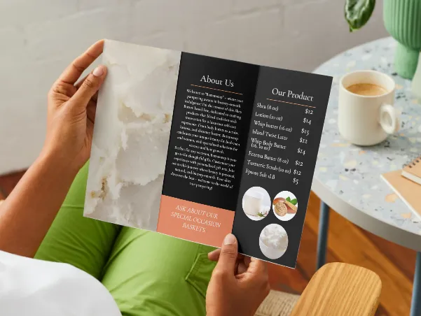 A person in green trousers holding open a shea butter tri-fold brochure beside a terrazzo side table with a coffee mug.