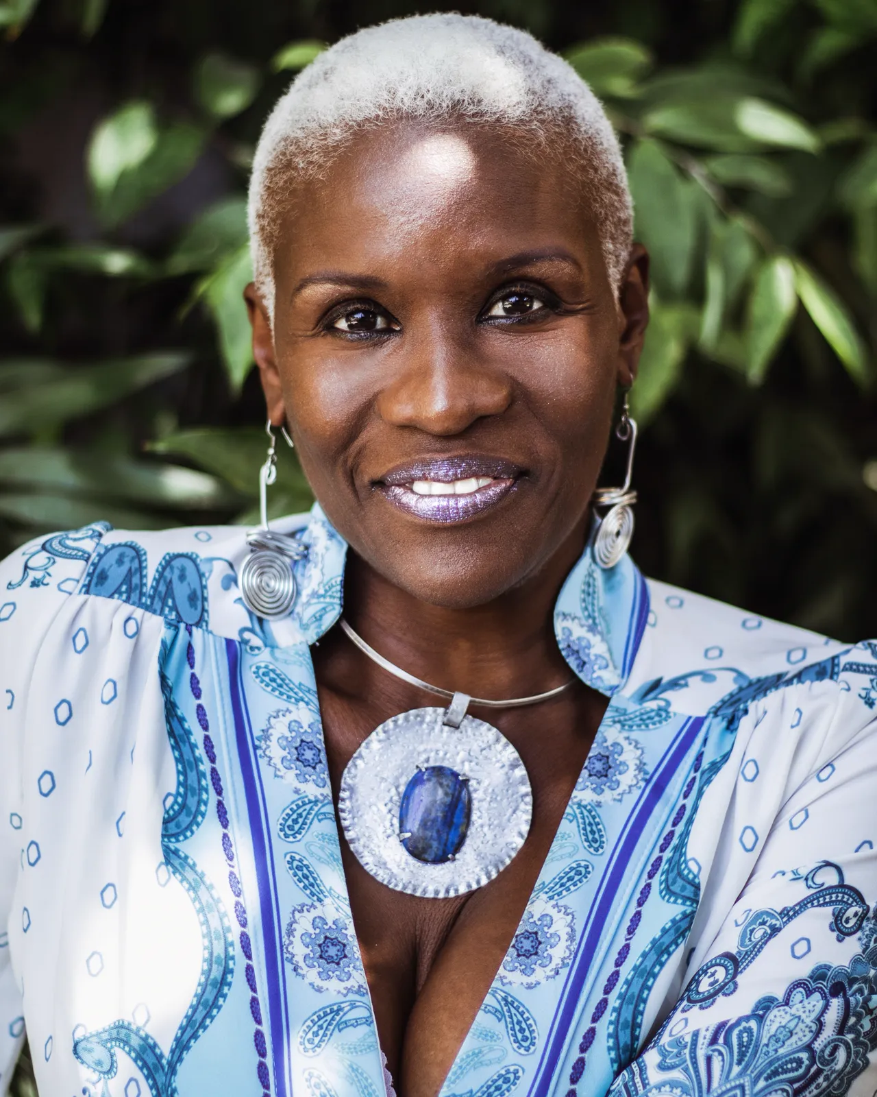 Close-up of a smiling woman with short platinum hair, silver earrings and a large blue stone pendant against green foliage.