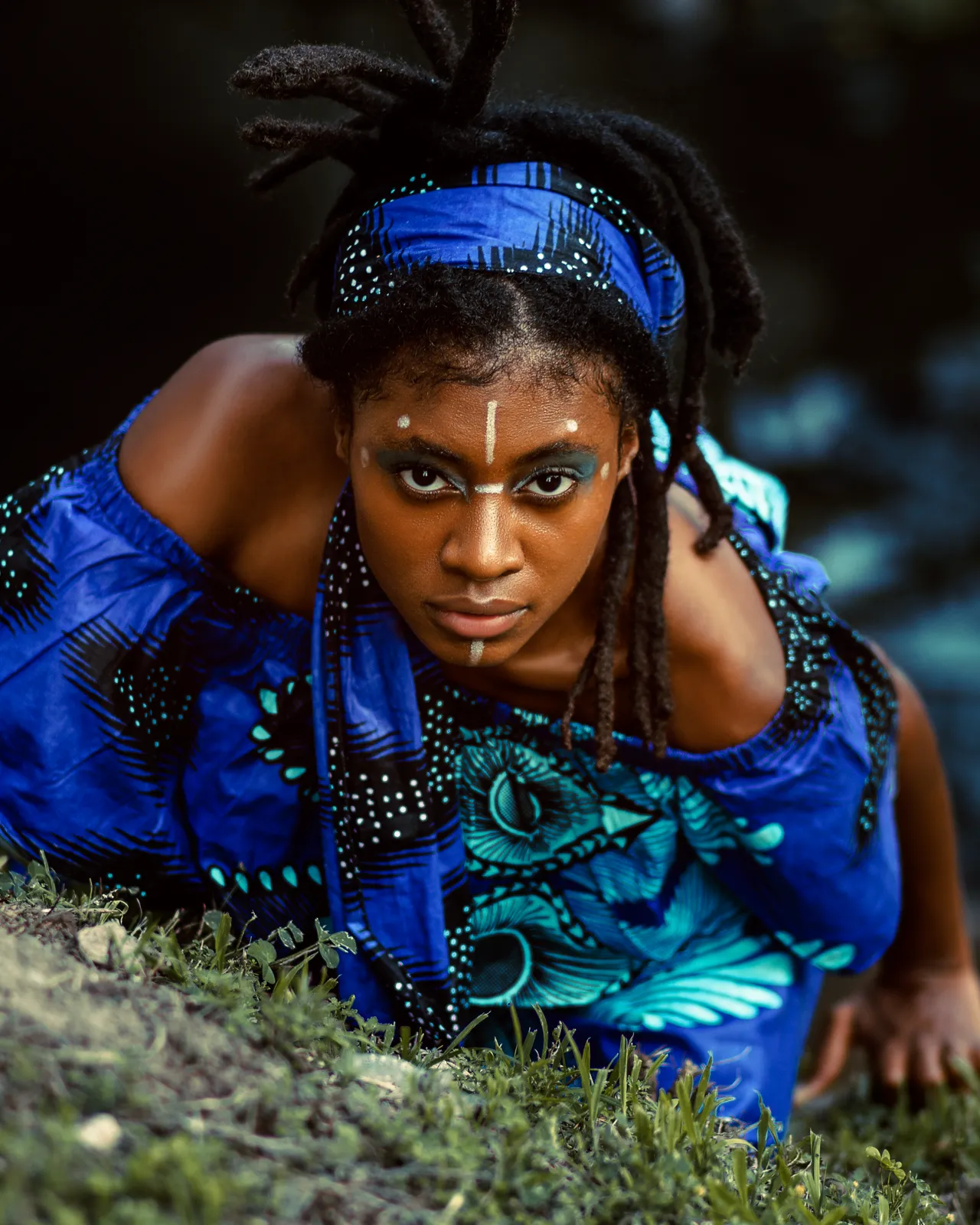 A woman in a blue African print off-shoulder dress and white face paint crouches on grass and stares intensely at the camera.