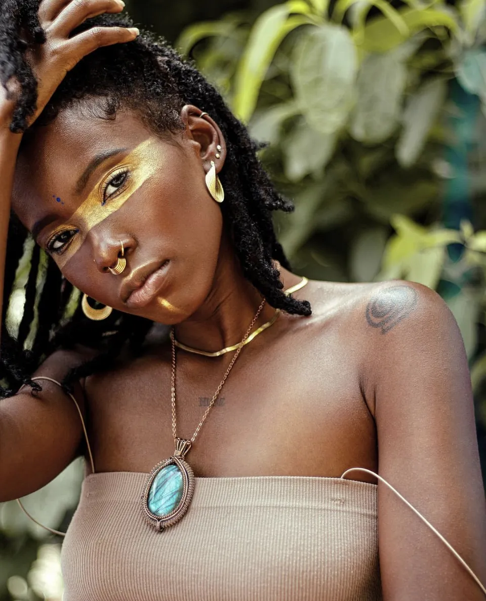 A woman with gold paint across her face and a turquoise pendant rests her hand on her head among leaves.
