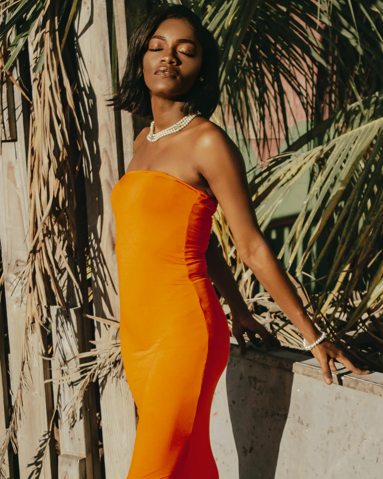 A woman in a fitted orange strapless dress poses with eyes closed beside a wall framed by palm fronds.