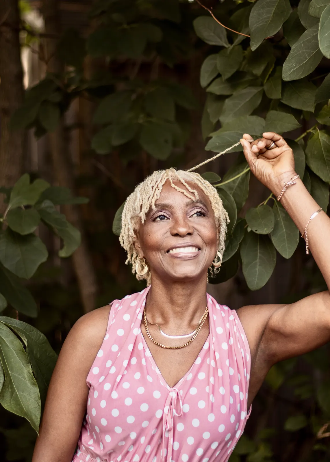 A smiling older woman with short blonde locs in a pink polka dot dress raises a hand to a leafy branch.