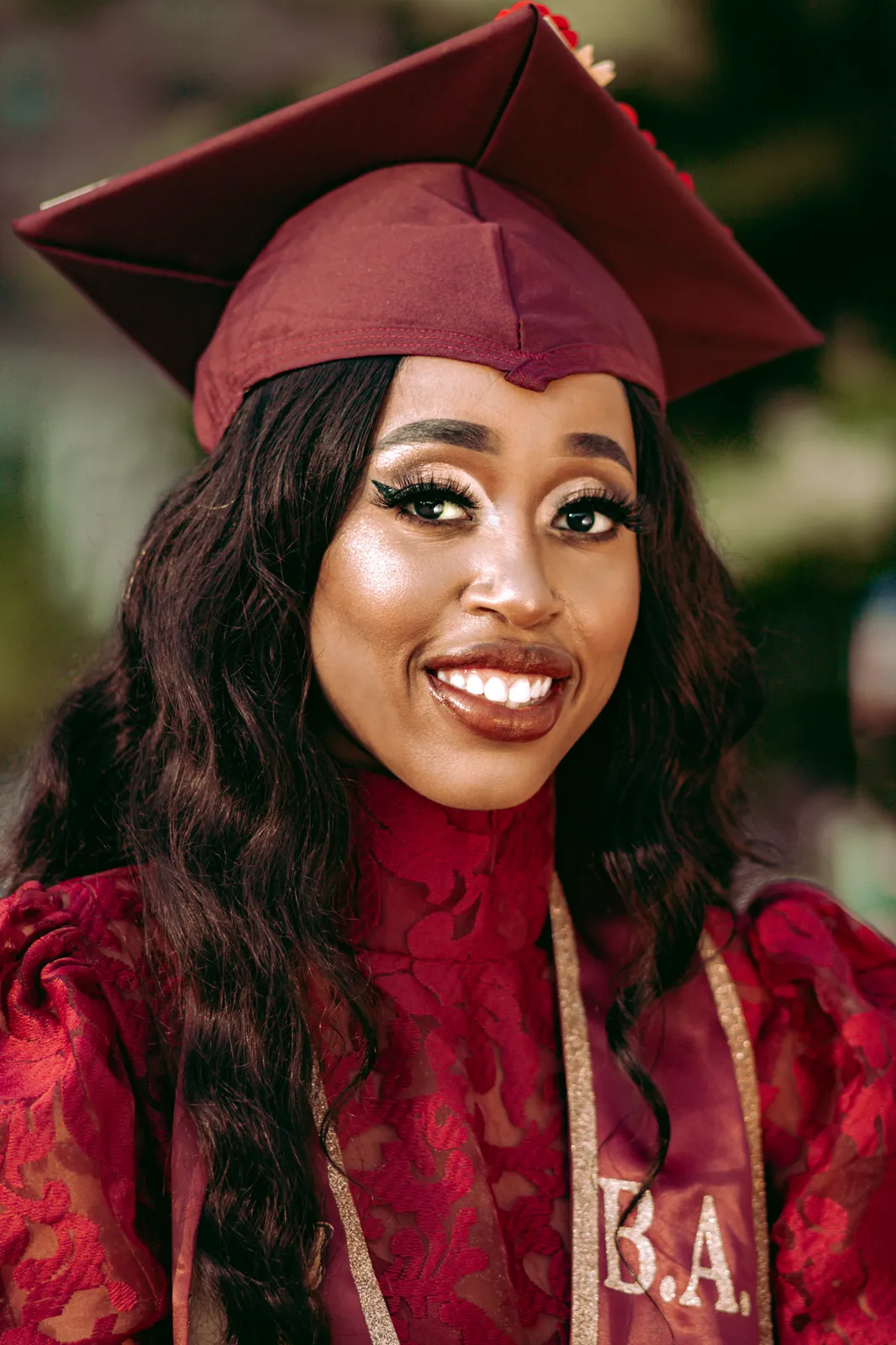A smiling graduate in a maroon cap, red lace dress and B.A. stole poses outdoors.