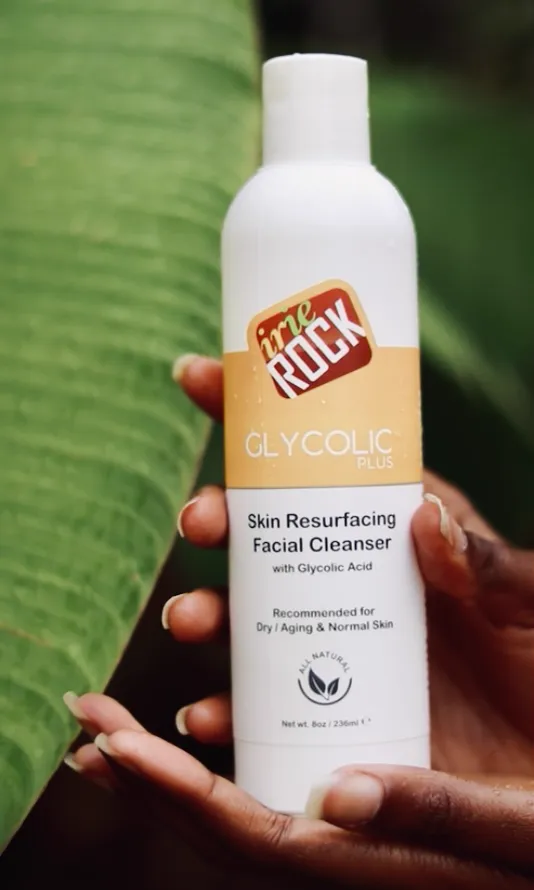 Hands hold an Irie Rock Glycolic Plus skin resurfacing facial cleanser bottle against a banana leaf.