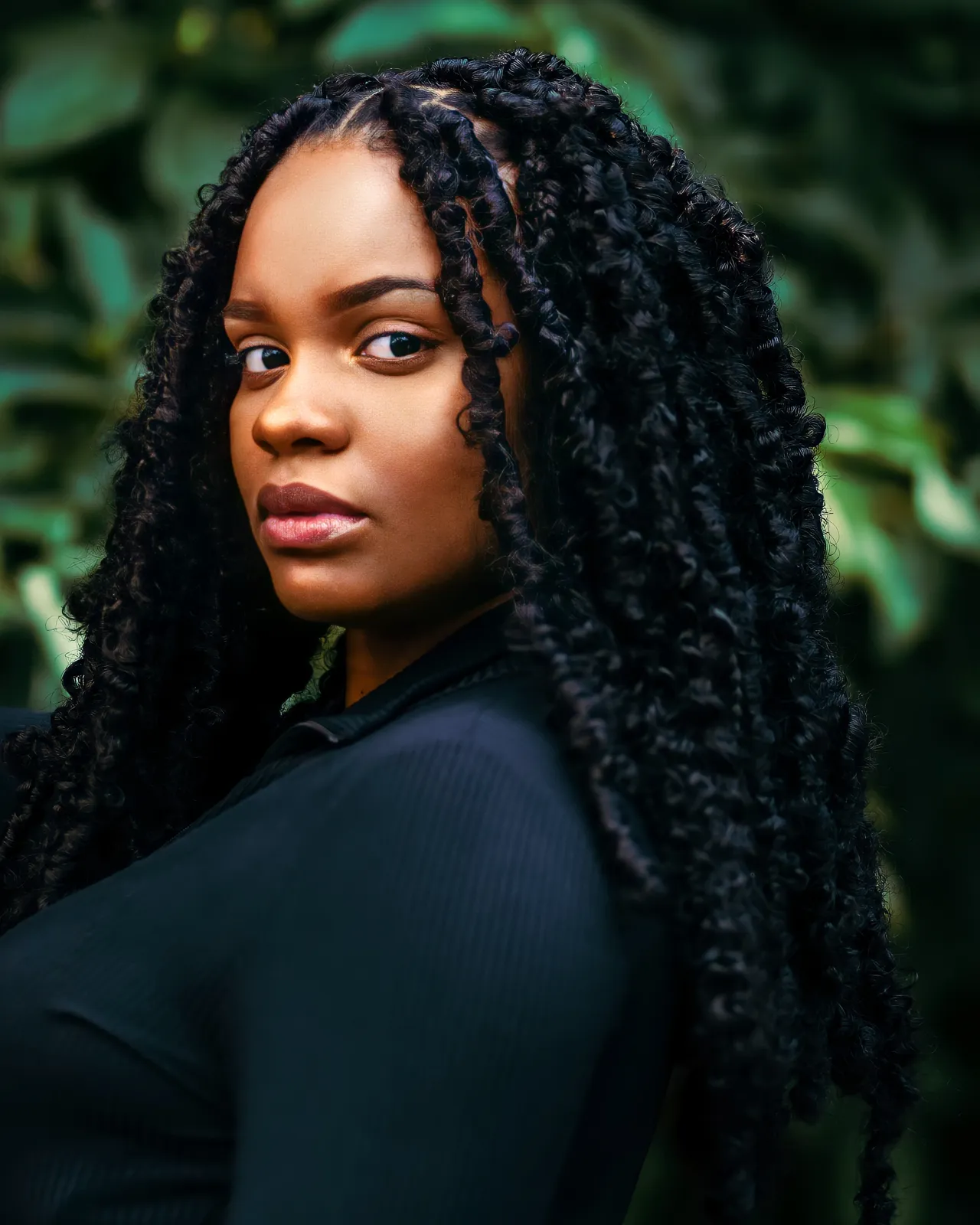 A woman with long black locs looks over her shoulder at the camera against dark green foliage.