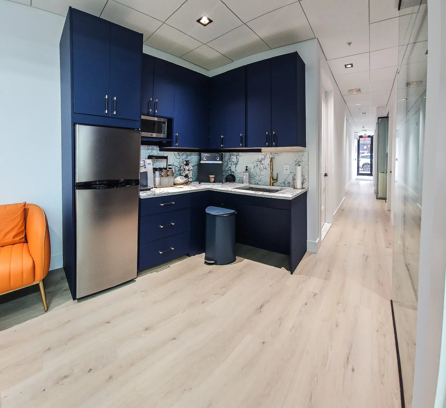 A modern kitchenette with navy cabinets, stainless refrigerator and light oak floors opens onto a long glass-walled hallway.