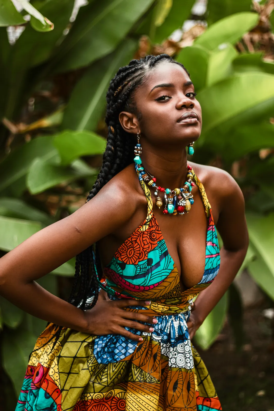 A woman with long braids in a colorful African print dress and beaded jewelry poses with hands on hips among tropical leaves.