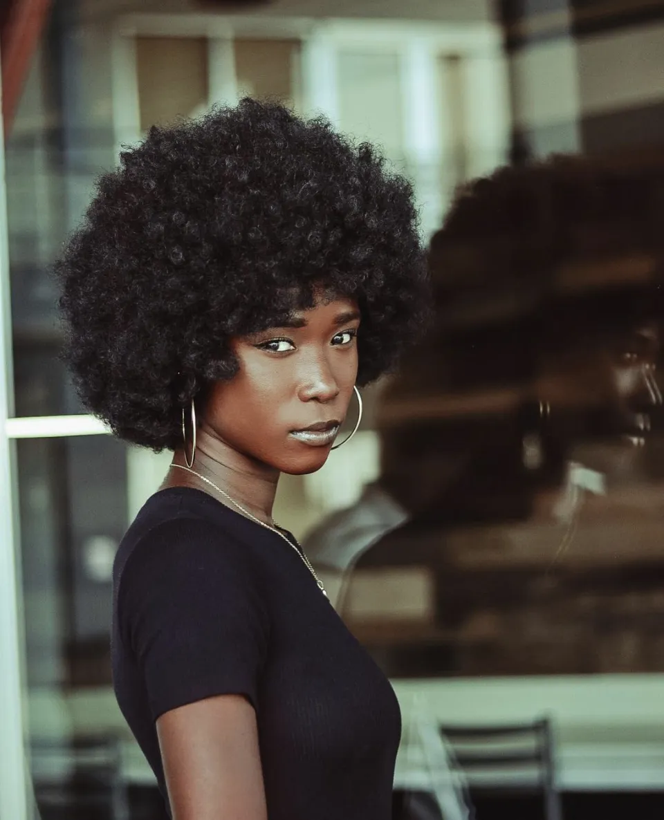 A woman with an afro, hoop earrings and a black top stands beside a window that reflects her profile.