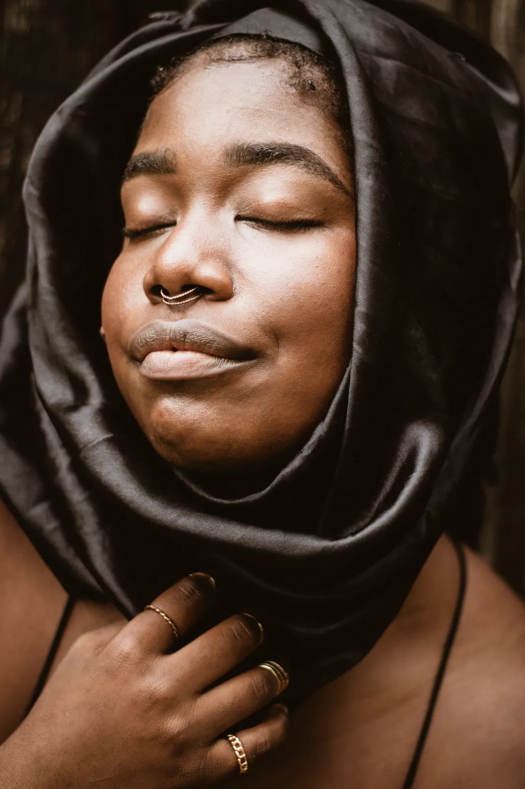 A woman wearing a black satin hood tilts her head back with eyes closed in warm light.
