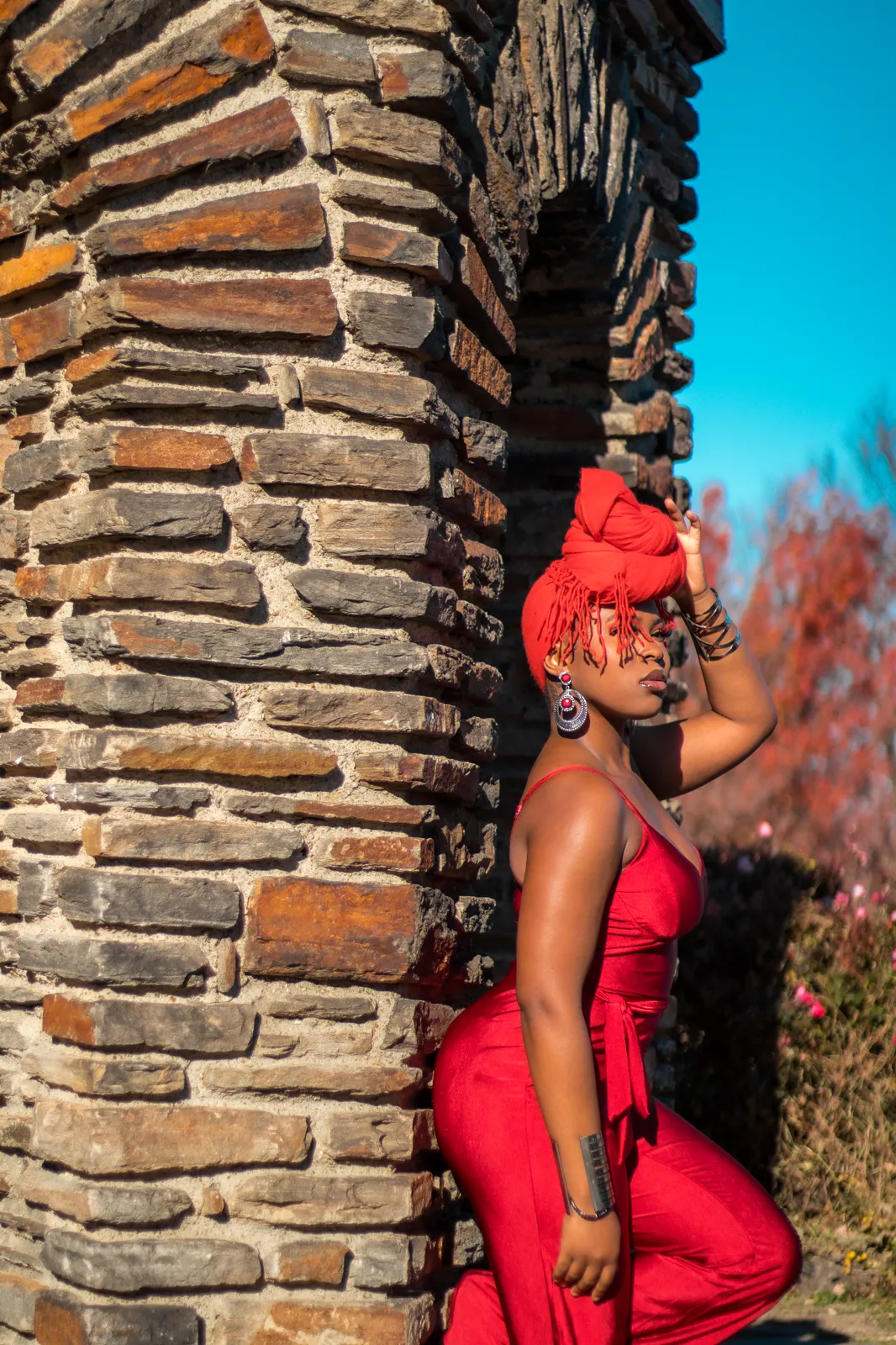 A woman in a red jumpsuit and head wrap leans against a stone pillar under a blue sky with red autumn foliage.