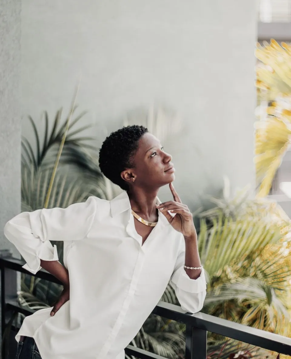 A woman with short hair in a white shirt leans on a balcony railing and gazes upward with palms behind.