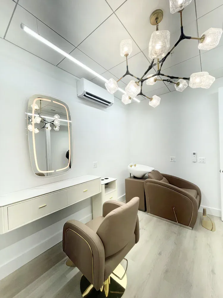 A white private salon suite with a branch chandelier, gold-framed mirror, taupe styling chair and shampoo station.