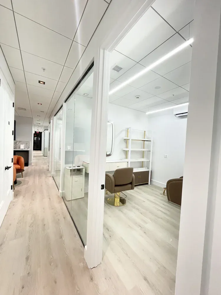 A bright corridor with glass-walled private salon rooms, light oak floors and linear ceiling lights.