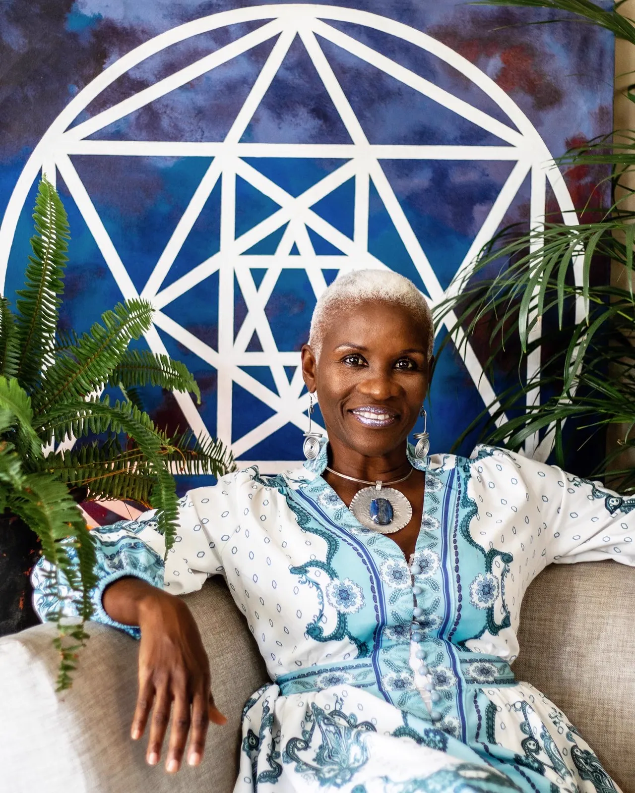 A smiling woman with short platinum hair in a blue and white paisley dress sits on a sofa in front of a blue geometric mural framed by ferns.