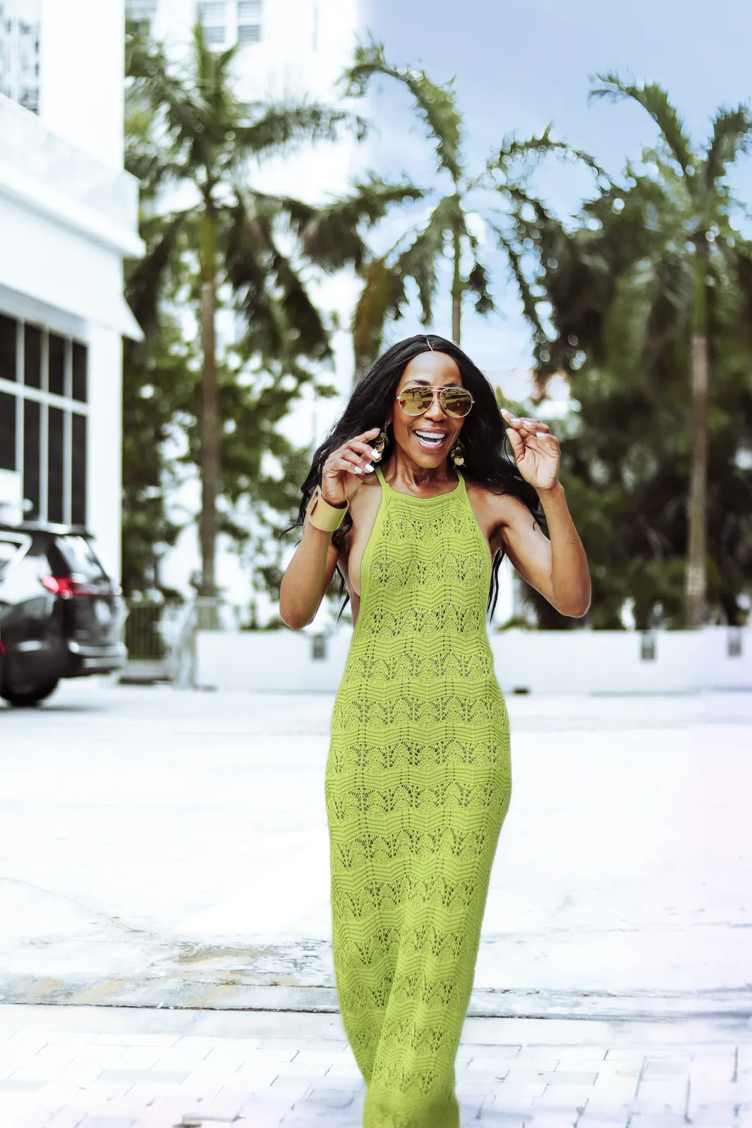 A smiling woman in sunglasses and a lime green knit dress walks a sunny street lined with palm trees.