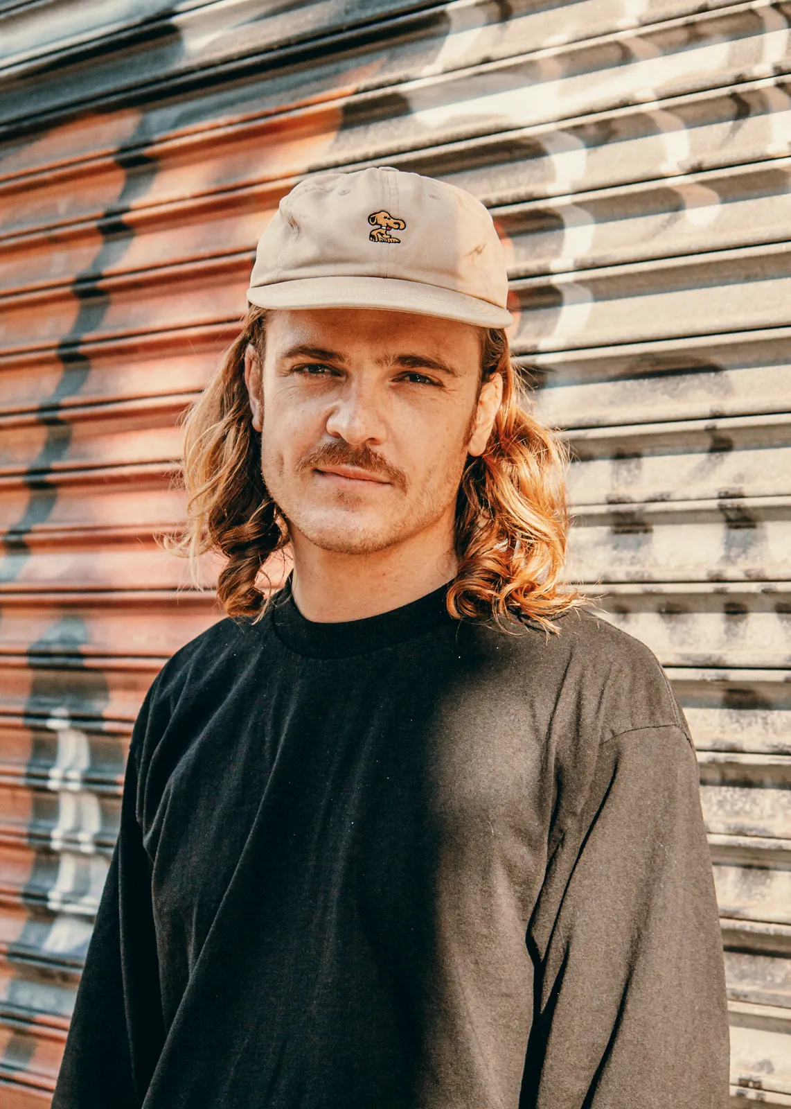 A man with long hair and a tan cap stands in front of a graffiti-marked metal shutter in split light.