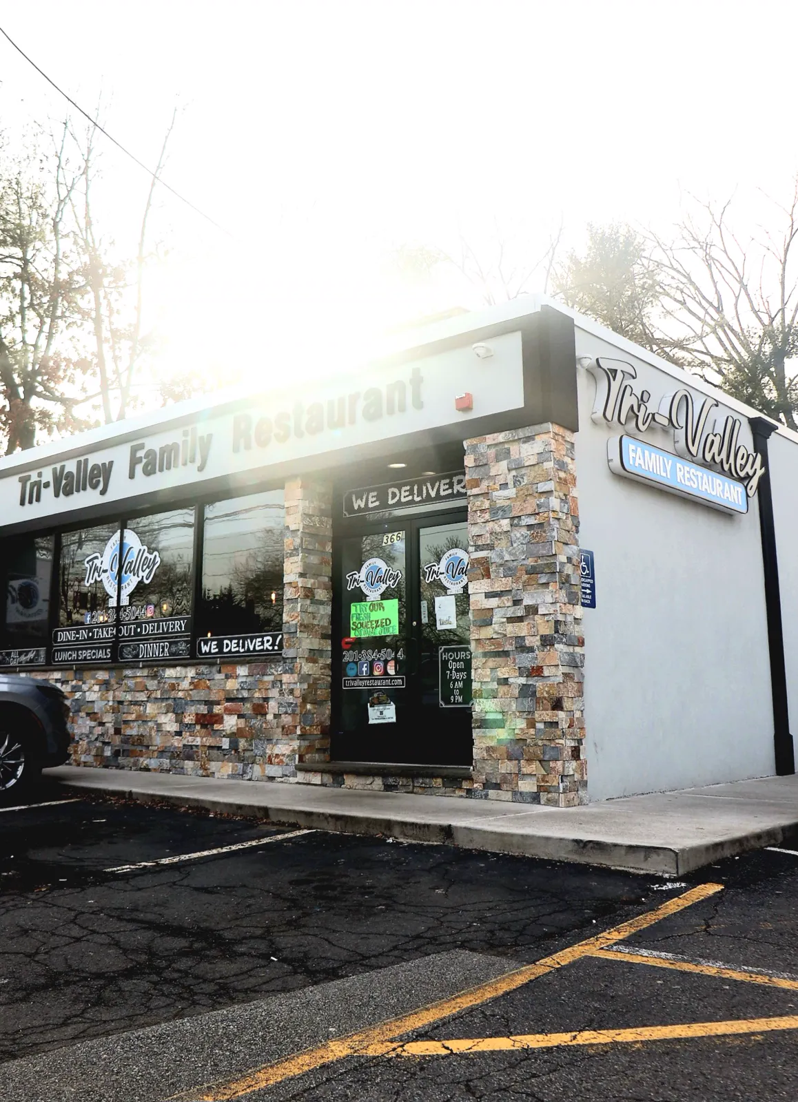 The stone-clad storefront of Tri-Valley Family Restaurant with We Deliver signage, lit by a bright sun flare.